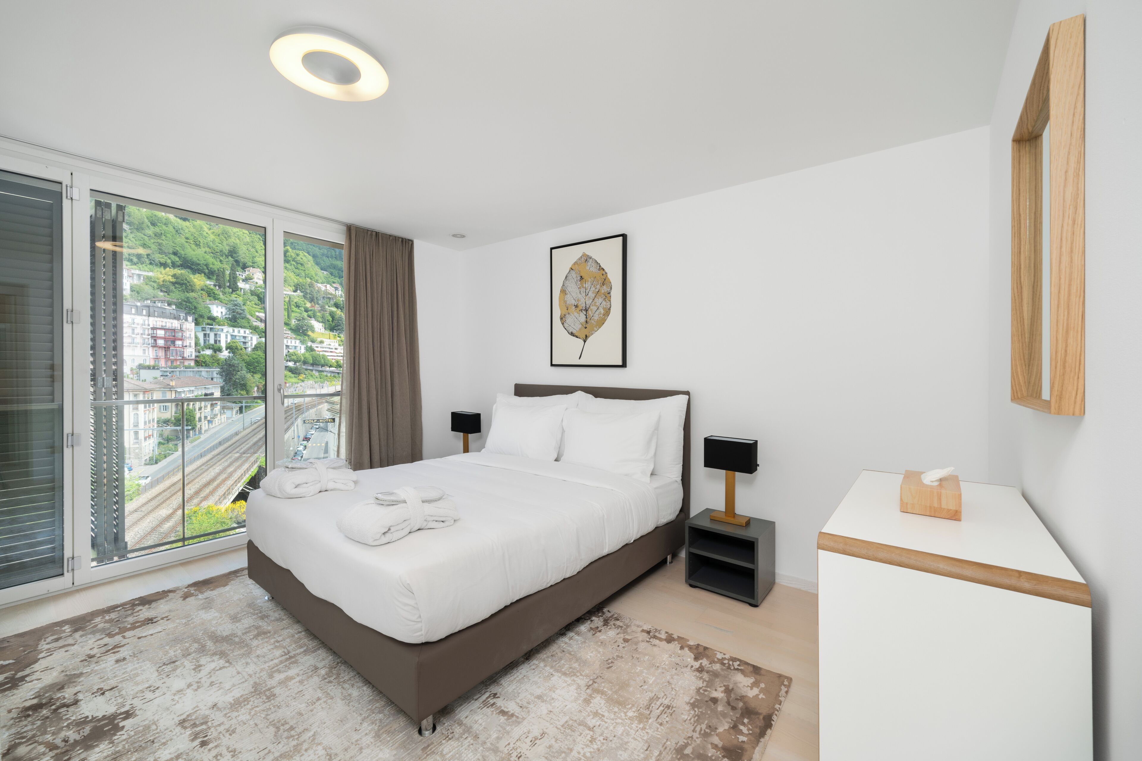 Une chambre d’hôtel moderne avec un grand lit, une table de chevet, une commode, une œuvre d’art encadrée, une fenêtre avec vue et un petit bureau avec une lampe.