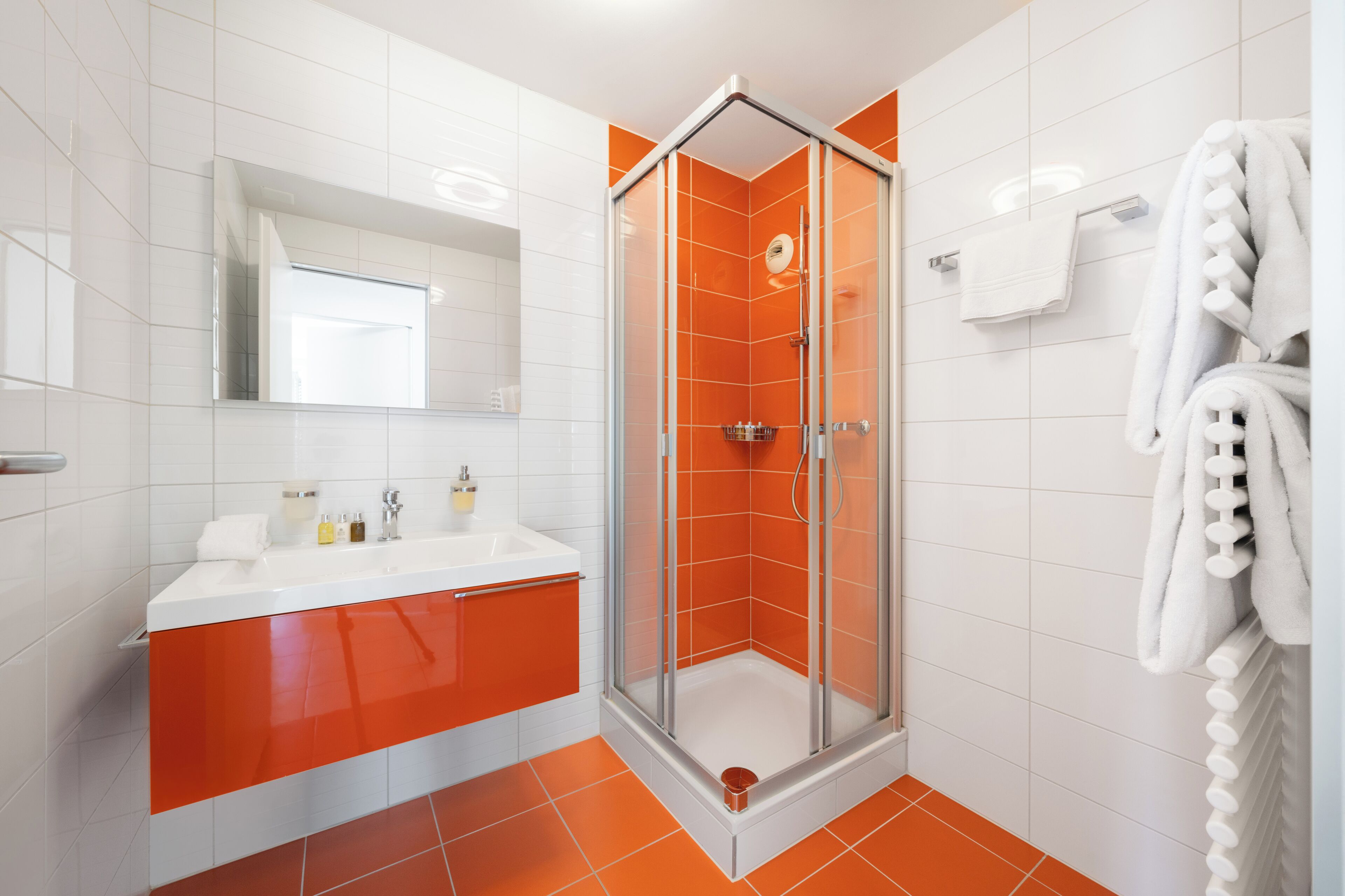 A modern bathroom with a glass-enclosed shower, white and orange tiles, a white sink with a mirror, and a towel rack with towels.
