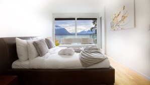 Een slaapkamer met een bed, kussens, een raam met uitzicht op bergen en water, en een wanddecoratie.