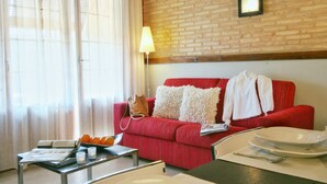 Un sofá rojo con almohadillas blancas, una mesa con lámpara y una ventana con cortinas traslúcidas.
