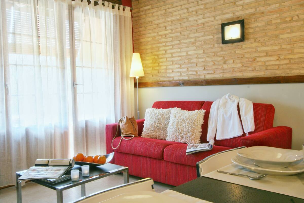 Un canapé rouge avec des coussins blancs, une table avec une lampe et une fenêtre ornée de rideaux légers.