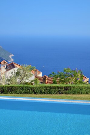 A swimming pool with a clear blue water surface, surrounded by a well-maintained hedge. In the background, there are buildings with red-tiled roofs and a view of the sea. - Villa Acacia (Kas)