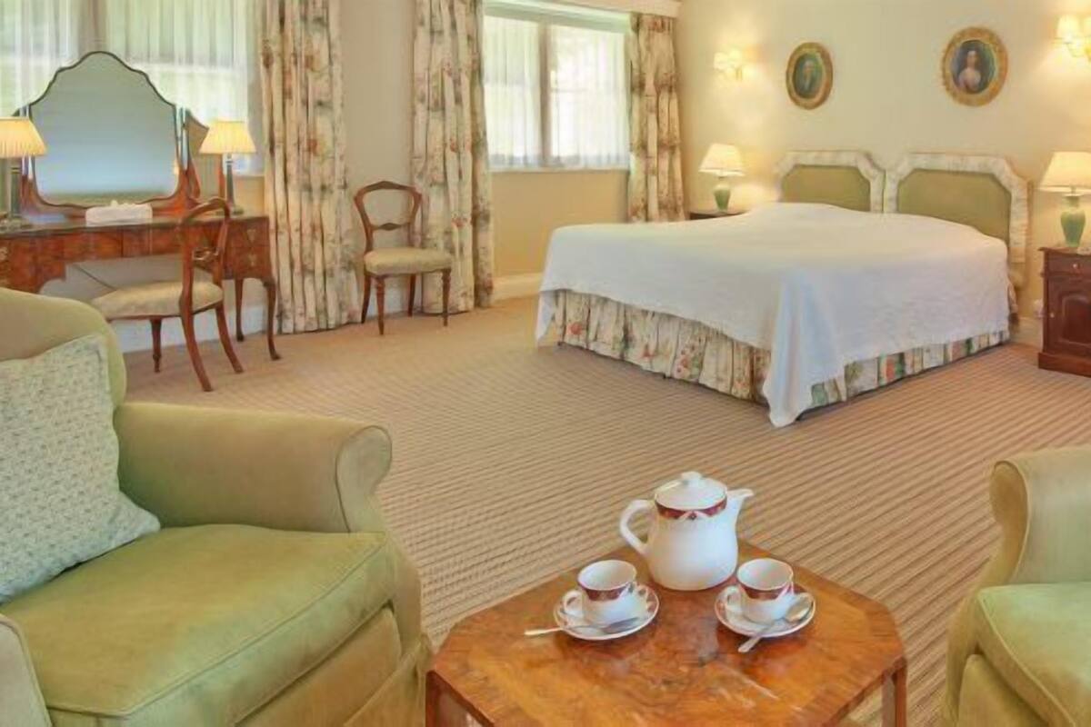 Ein Hotelzimmer mit einem Bett, zwei Sesseln, einem Holztisch mit Teeservice, einem Schreibtisch mit Spiegel und zwei Wandleuchten.