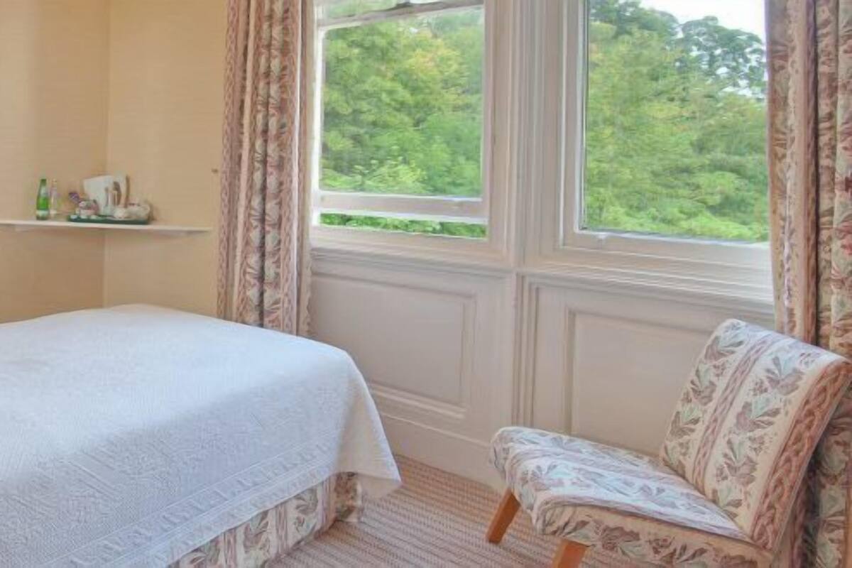 Ein Schlafzimmer mit einem Bett, einem Sessel, einem Nachttisch, einer Lampe, einem Regal mit Pflegeprodukten und einem Fenster mit Blick ins Grüne.