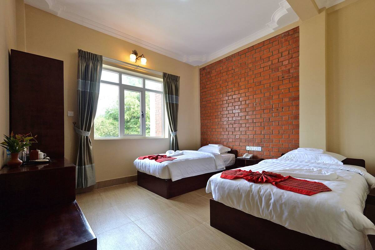 A hotel room with two beds, a brick wall, a window with curtains, and a desk with a plant.