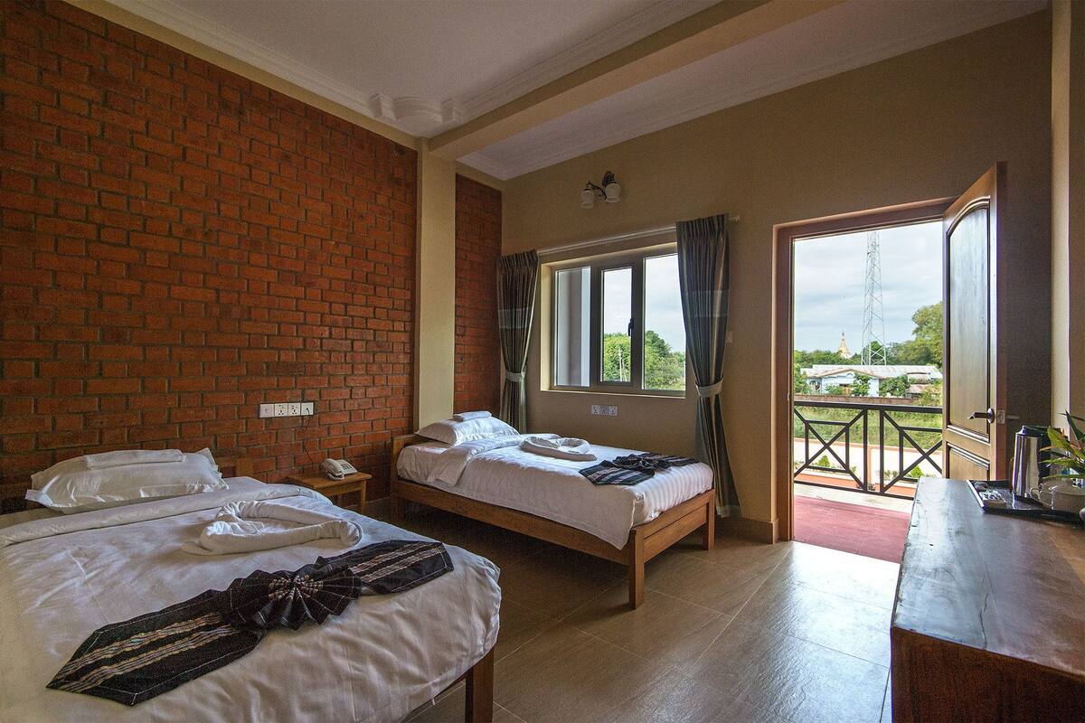 A hotel room with two beds, a brick wall, a window with curtains, and a balcony with a view of greenery and buildings.