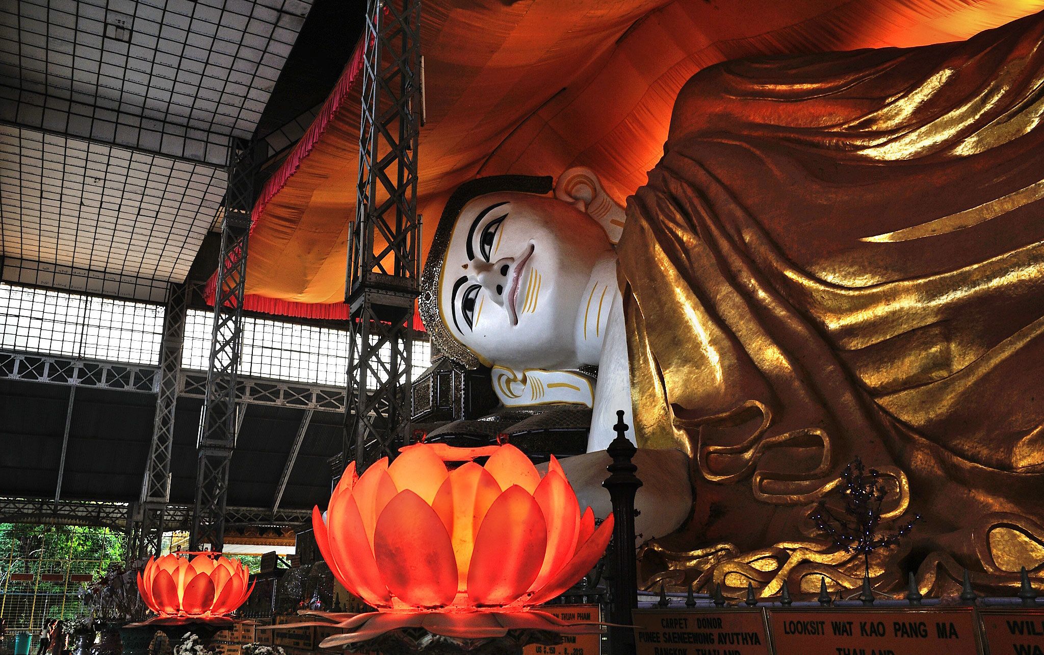 Una gran estatua de Buda con túnica dorada y rostro blanco, rodeada por una lámpara de loto rojo y una estructura metálica.