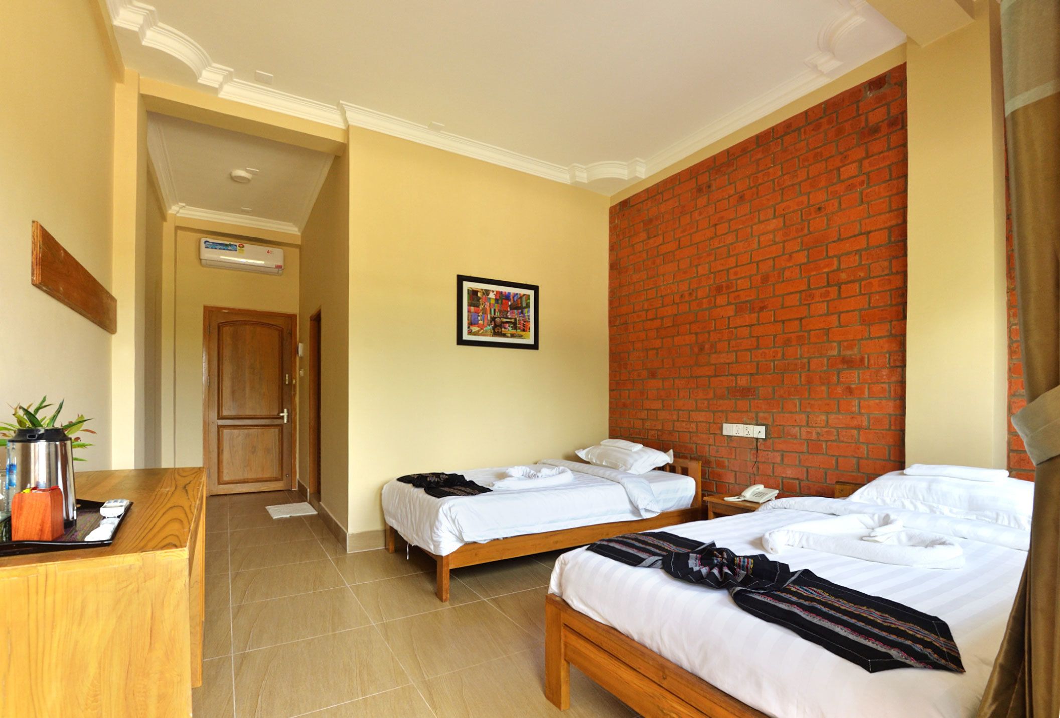 A hotel room with two beds, a wooden desk, a brick accent wall, and a framed picture on the wall.