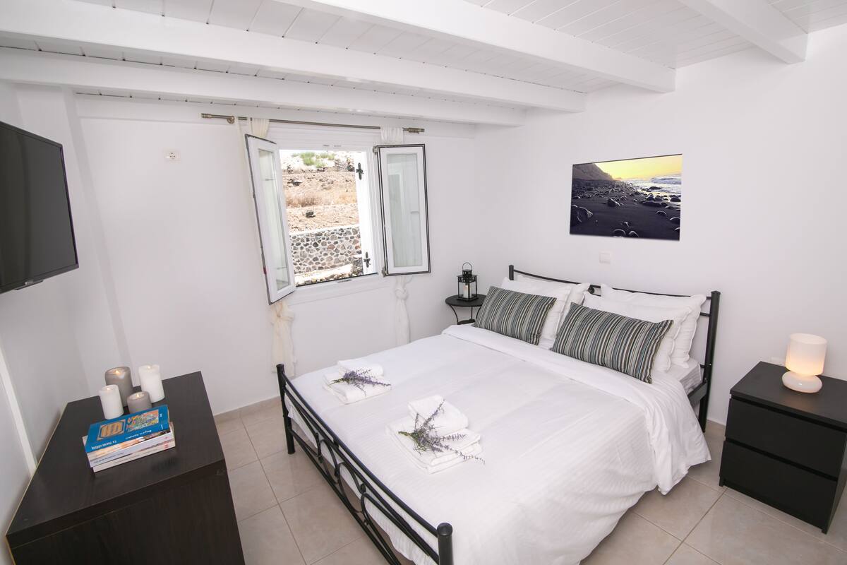 Ein ordentlich eingerichtetes Schlafzimmer mit Bett, Nachttischen, einem Wand-Fernseher, einem Fenster mit geöffnetem Vorhang und einem gerahmten Bild an der Wand.