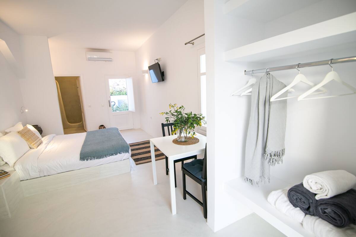 Ein modernes Hotelzimmer mit einem Bett, einem Esstisch mit Stühlen, einer kleinen Pflanze, einem an der Wand montierten Fernseher und einem Kleiderständer mit Kleiderbügeln.