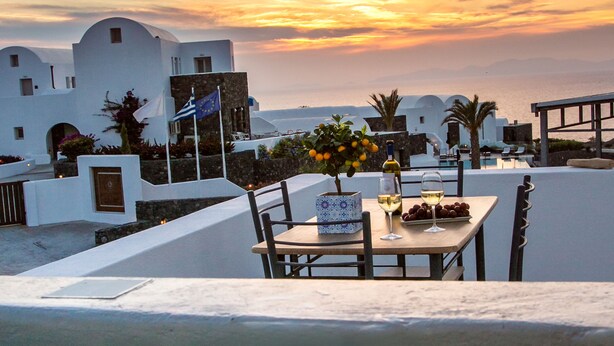 A table set with a bottle of wine, glasses, and a plate of olives, overlooking a coastal area with white buildings and a sunset. - Apanomeria Residence (Santorini)