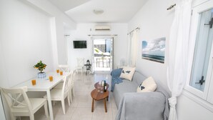 A bright, white room with a dining table, chairs, a sofa, and a TV on the wall. - Apanomeria Residence (Santorini)