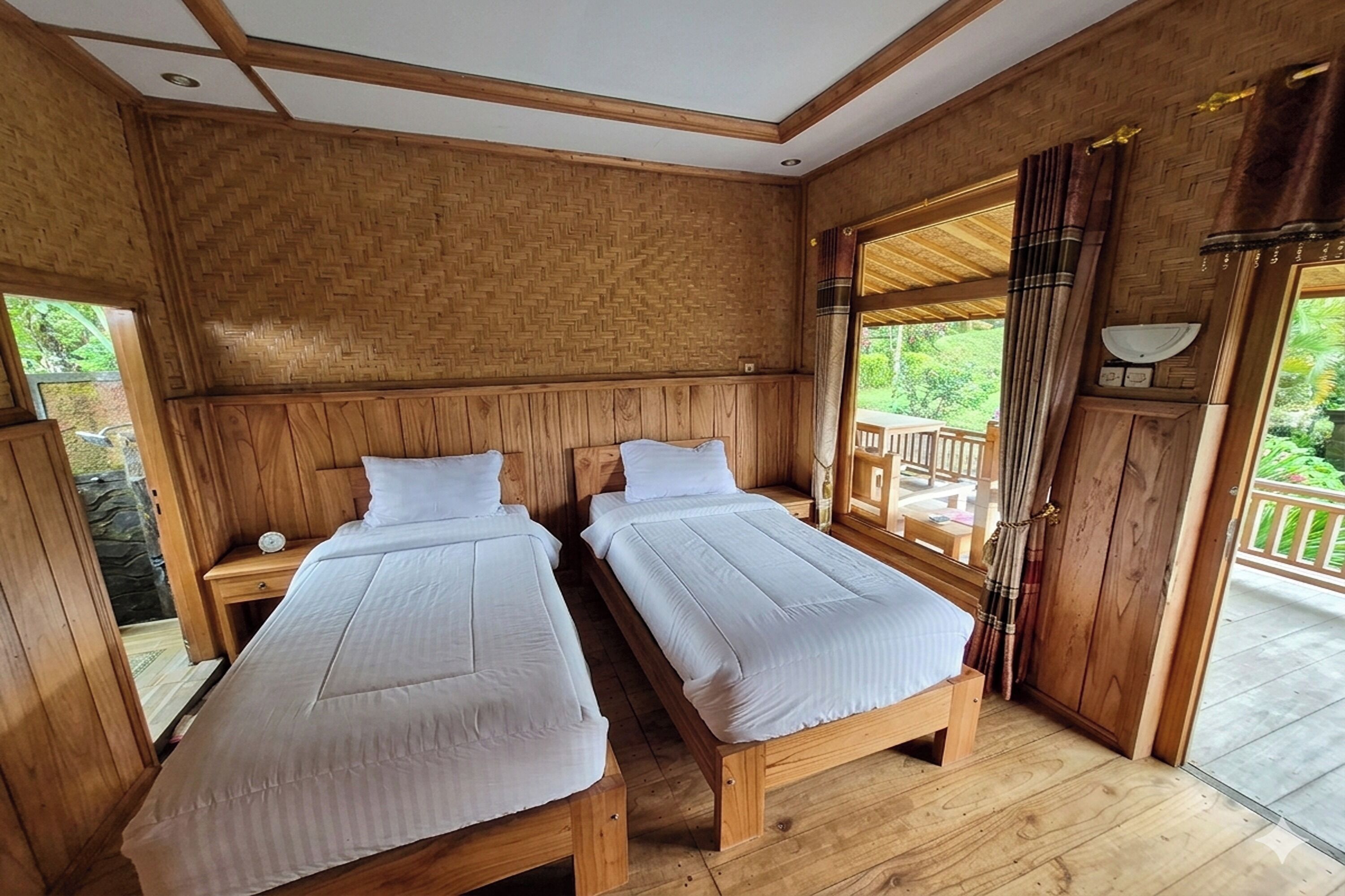 A wooden room with two beds, a nightstand, a window with curtains, and a view of greenery outside.