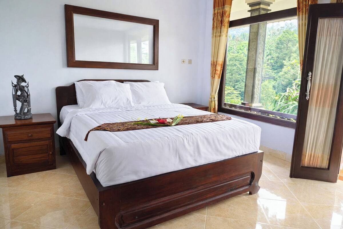 A bedroom with a bed, a nightstand, a mirror, and a window with a view of greenery.