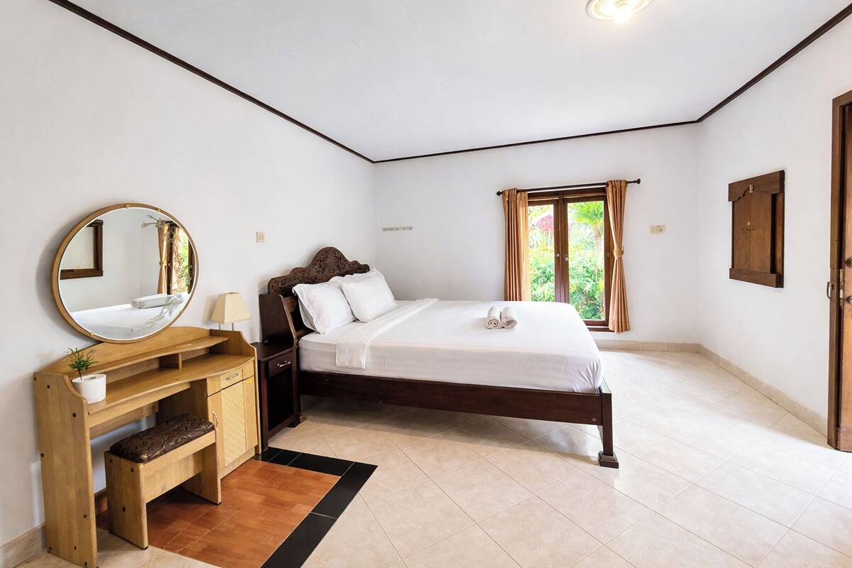 A bedroom with a bed, a wooden desk with a mirror, a small bench, and a window with curtains.