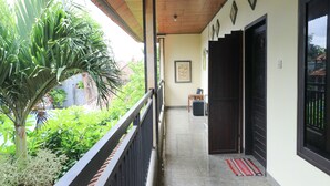 A covered balcony with a view of lush greenery, a wooden ceiling, and a door leading to a room. - Kubu Turah Homestay (Kedonganan)