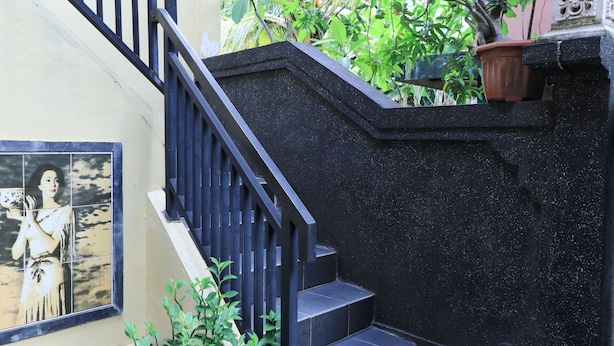 A staircase with black railings leading to a landing, surrounded by greenery and a tiled wall. - Kubu Turah Homestay (Kedonganan)