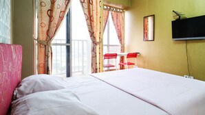 Habitación de hotel con cama, televisión montada en la pared y balcón con sillas rojas.