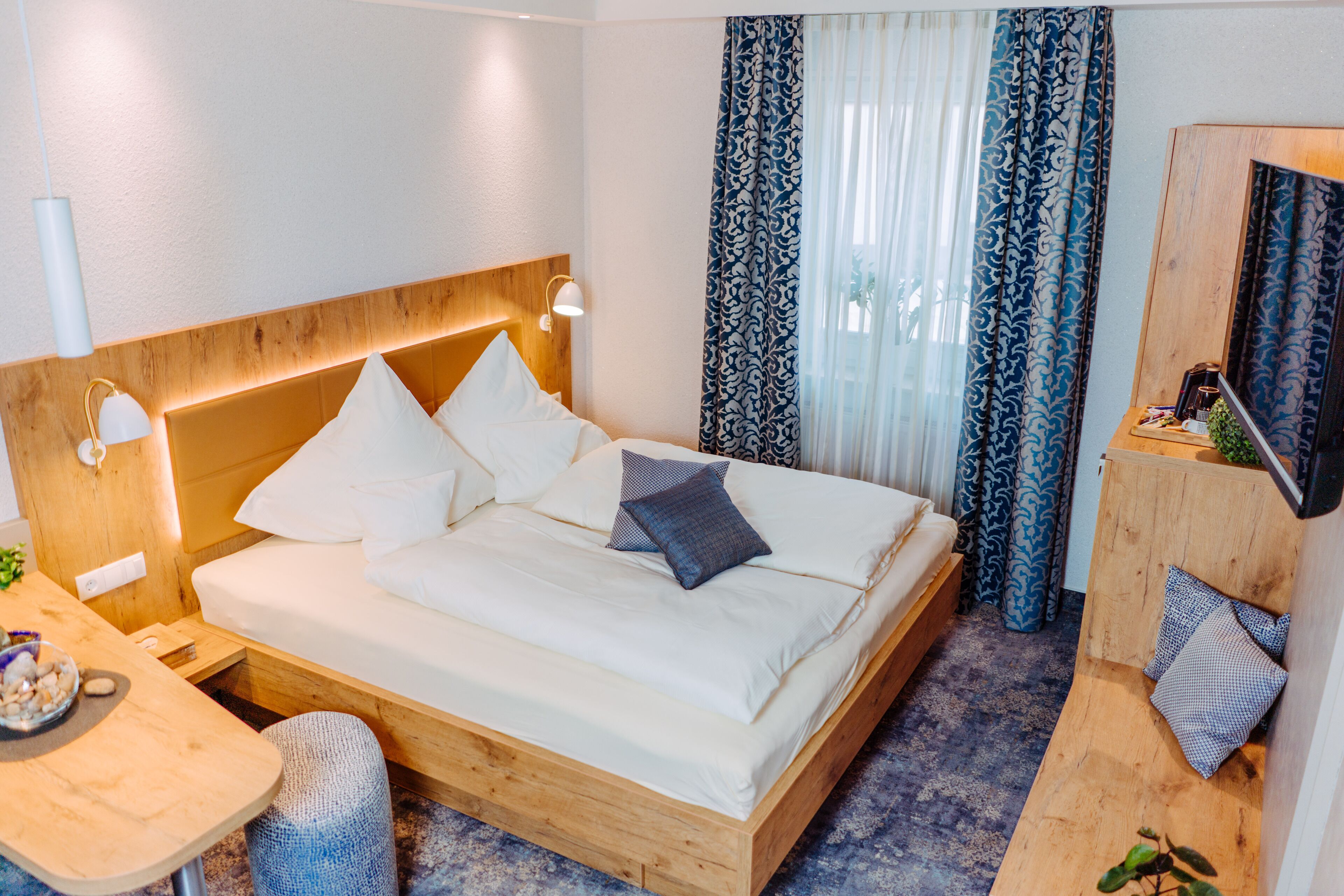 Habitación de hotel con cabecera de madera, cama con sábanas y almohadas blancas, una mesita con frutero, un banco de madera y un televisor sobre un mueble de madera.