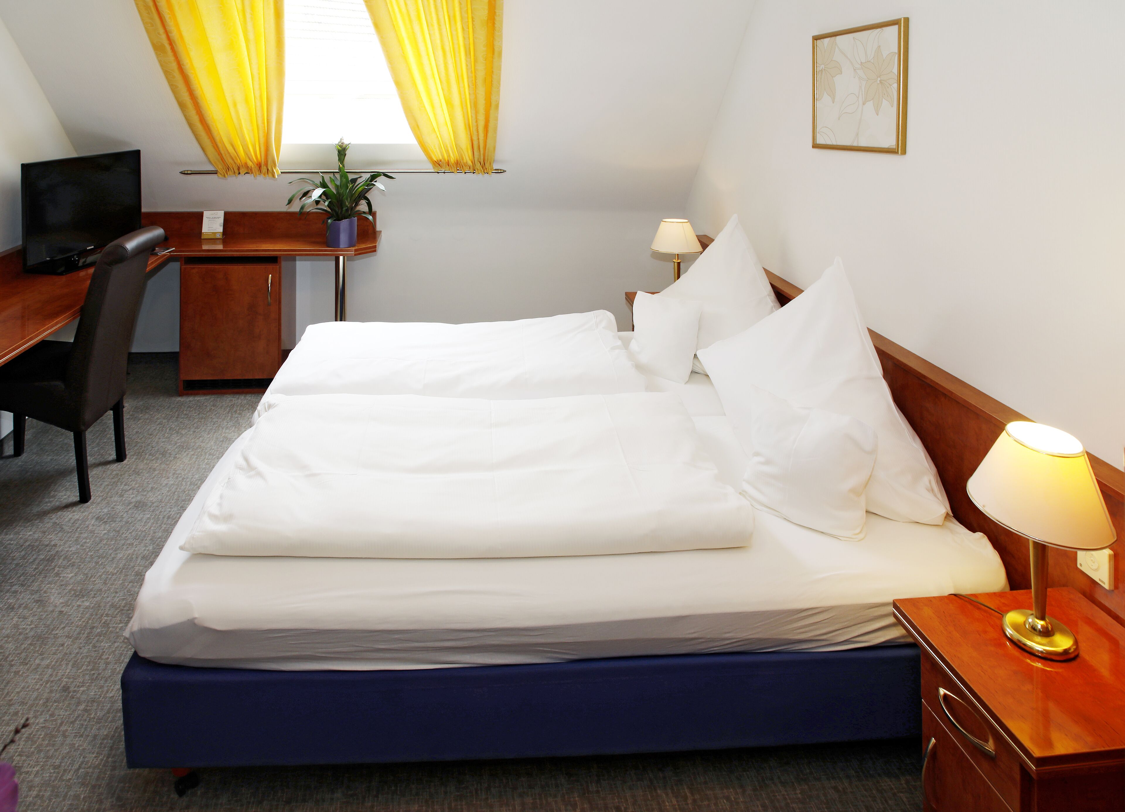 A hotel room with a large bed, a desk with a chair, a TV, a lamp, and a potted plant.