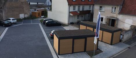 A street view with a row of parked cars, a building with a sloped roof, and a small structure with a flag.