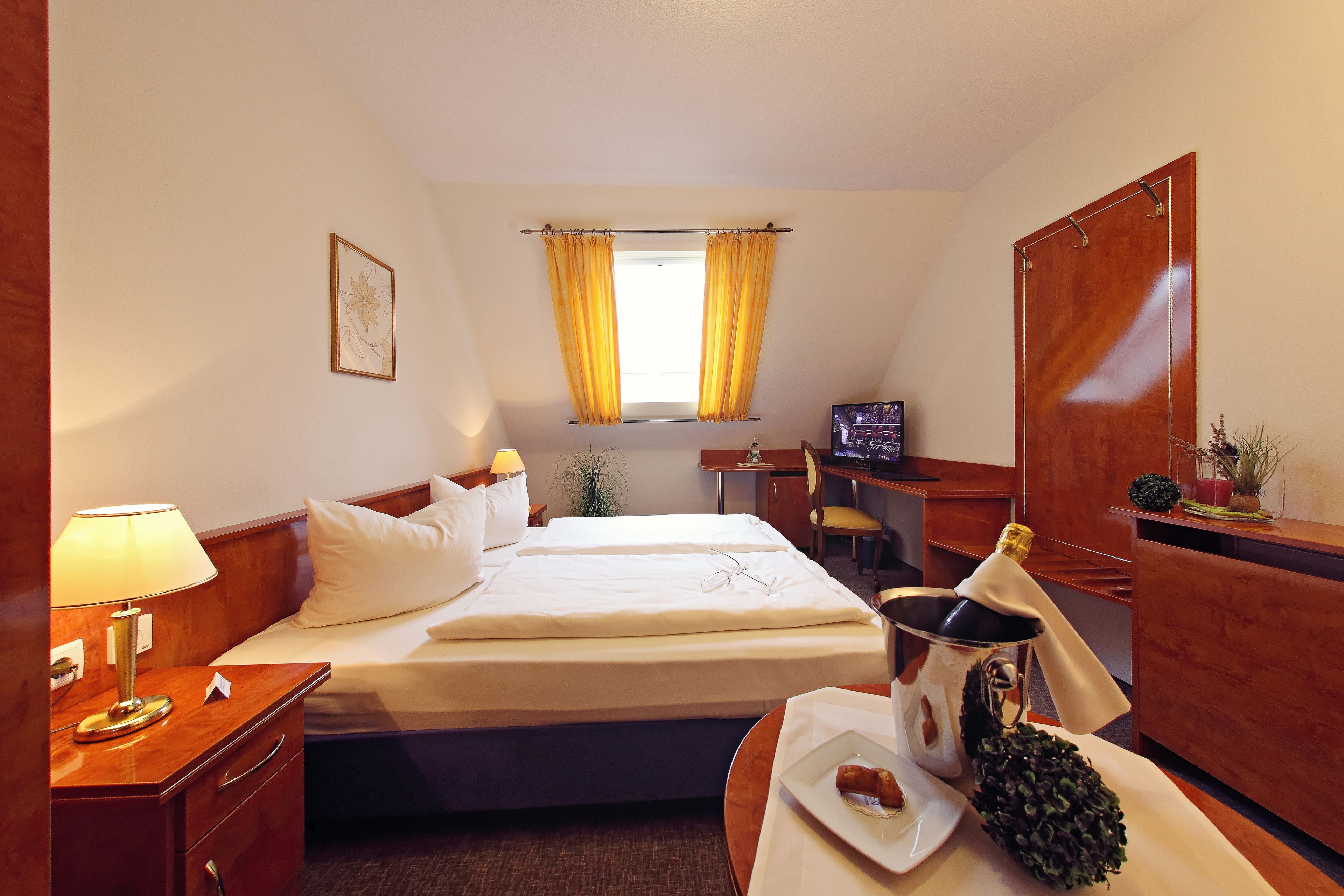 Habitación de hotel con cama, mesita de noche, lámpara, escritorio, silla y un ventanal con cortinas.