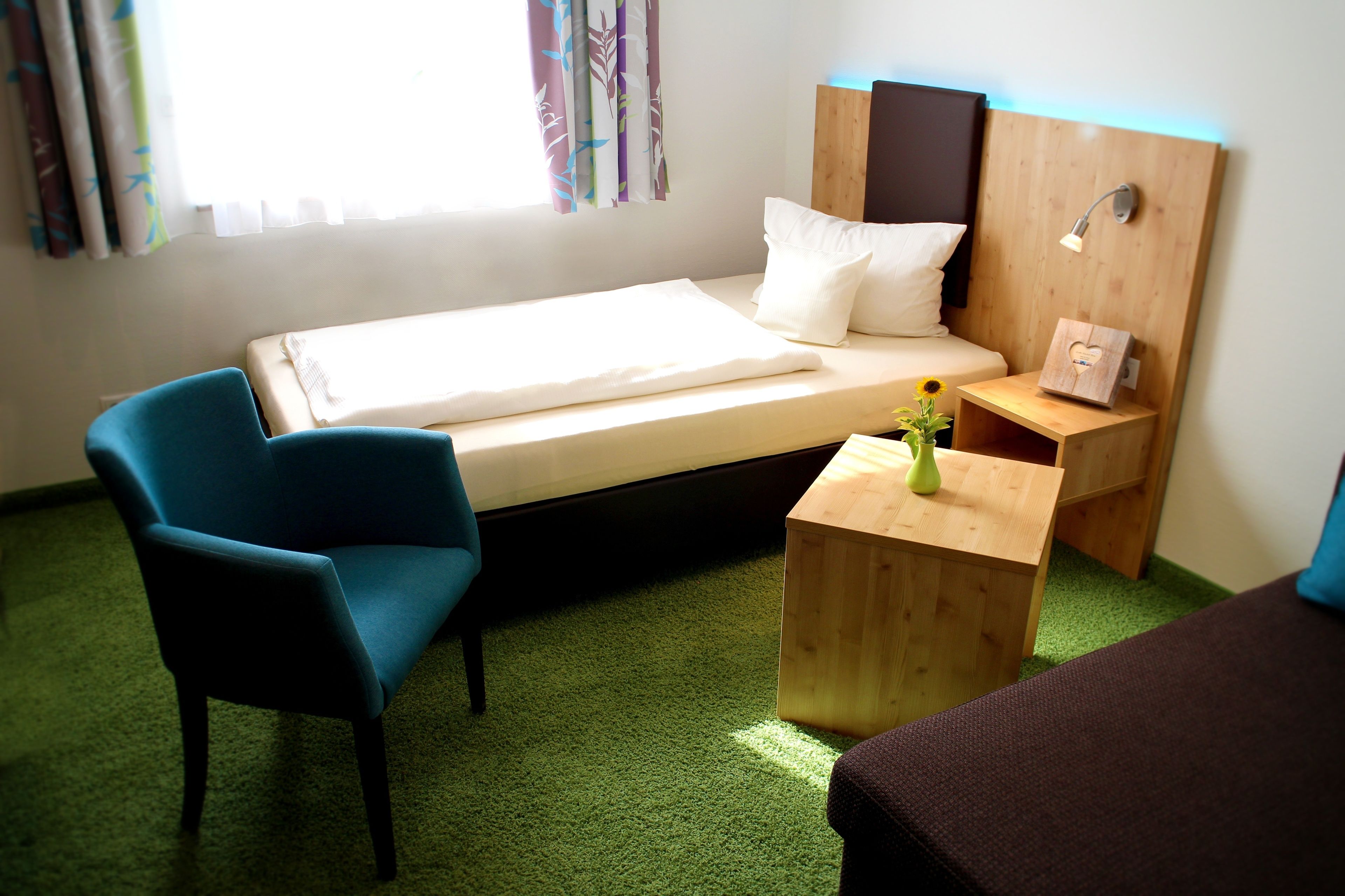 Habitación de hotel con una cama individual, una silla azul, una mesita de noche de madera con una lámpara y una pequeña mesa con un florero.
