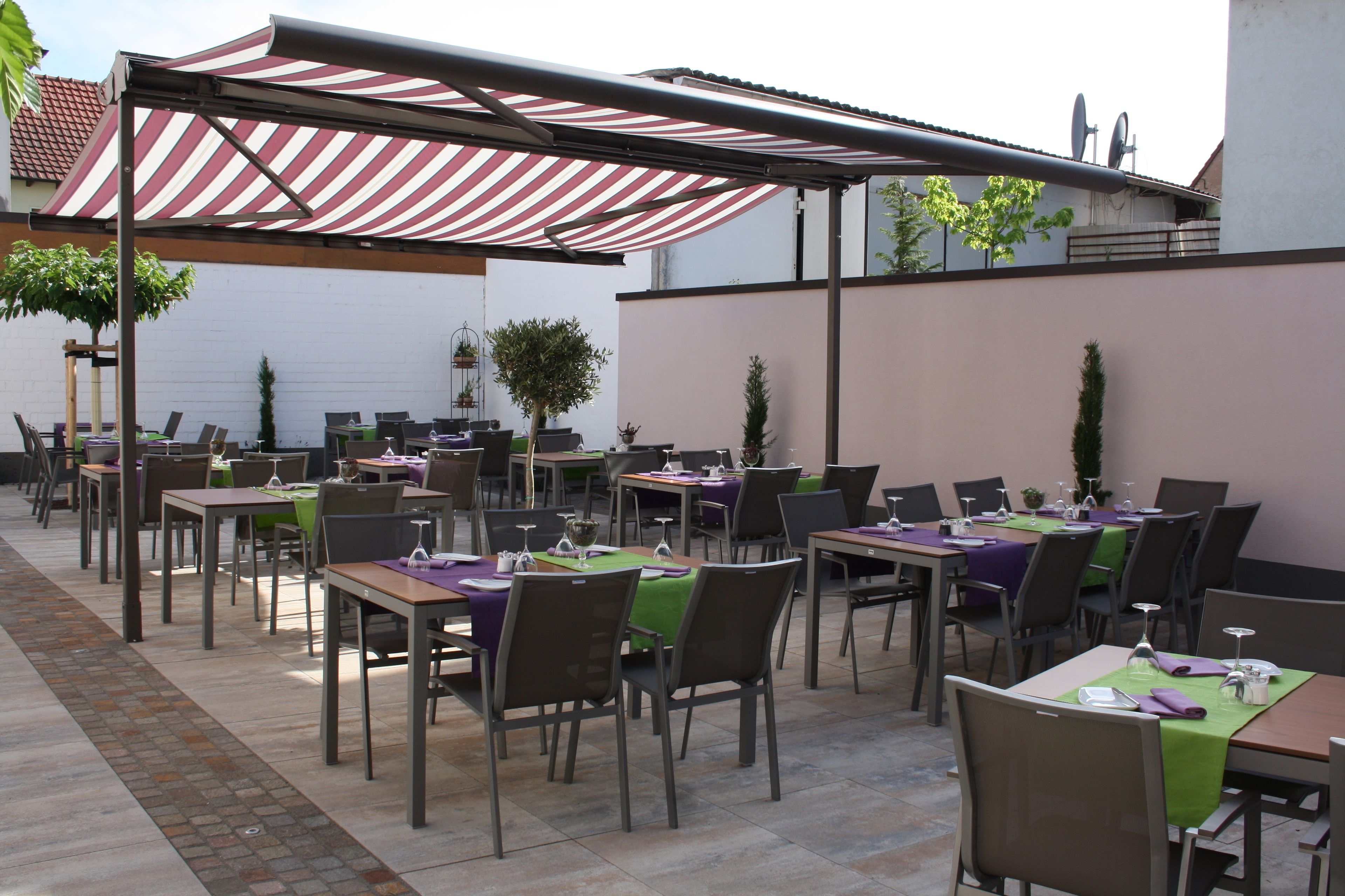 Un espace extérieur aménagé pour les repas, avec des tables et des chaises, protégé par des auvents à rayures et entouré de plantes en pot.