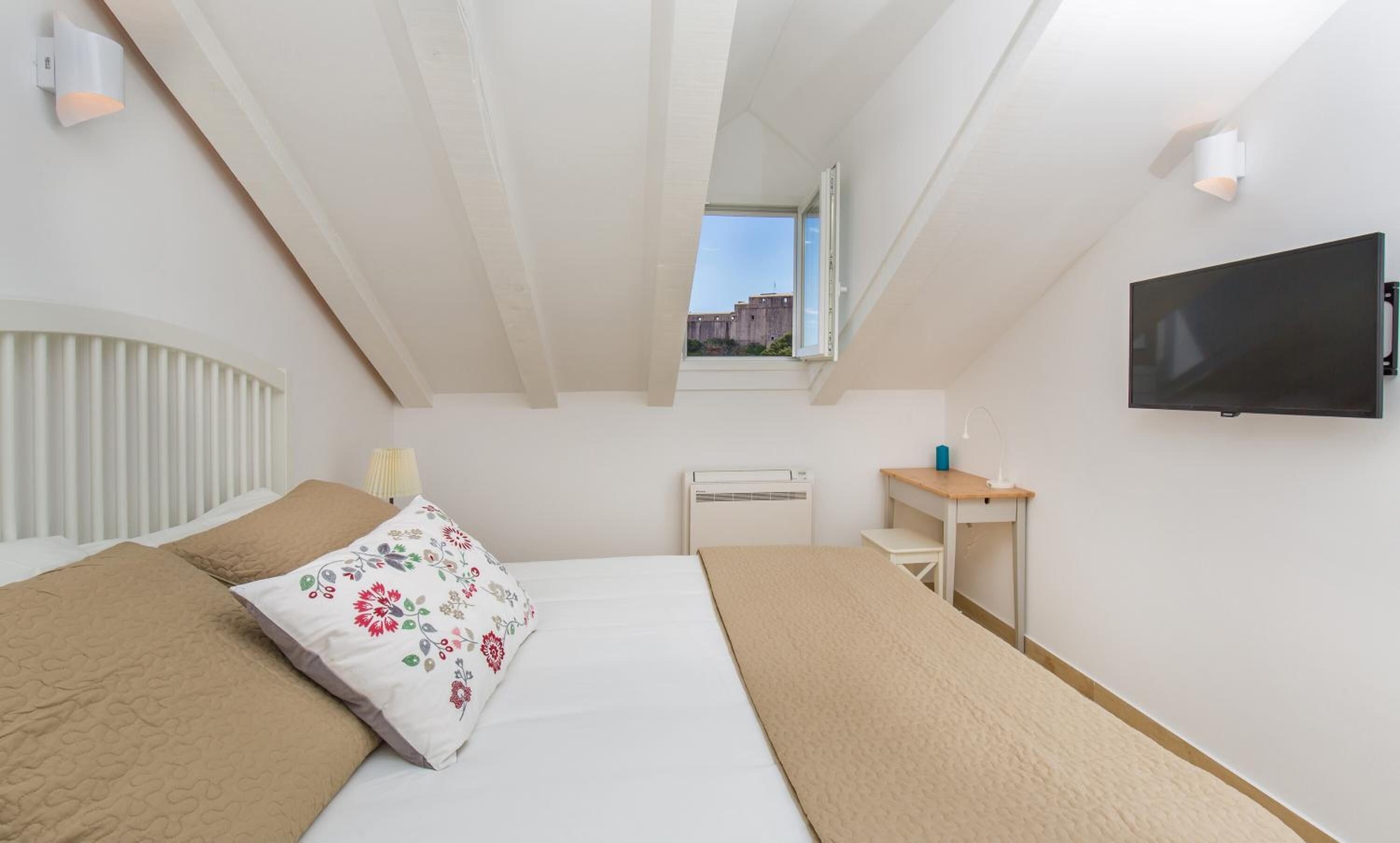 Ein Schlafzimmer mit Bett, Schreibtisch, Fernseher und einem Fenster mit Blick auf Gebäude.