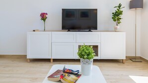 Ein modernes Wohnzimmer mit einem Flachbildfernseher auf einem weißen Schrank, einem Couchtisch mit Büchern und einer Zimmerpflanze sowie einer Stehlampe.
