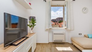 Ein modernes Wohnzimmer mit Flachbildfernseher, Zimmerpflanze, Fenster mit Vorhängen, Sofa und Heizkörper.