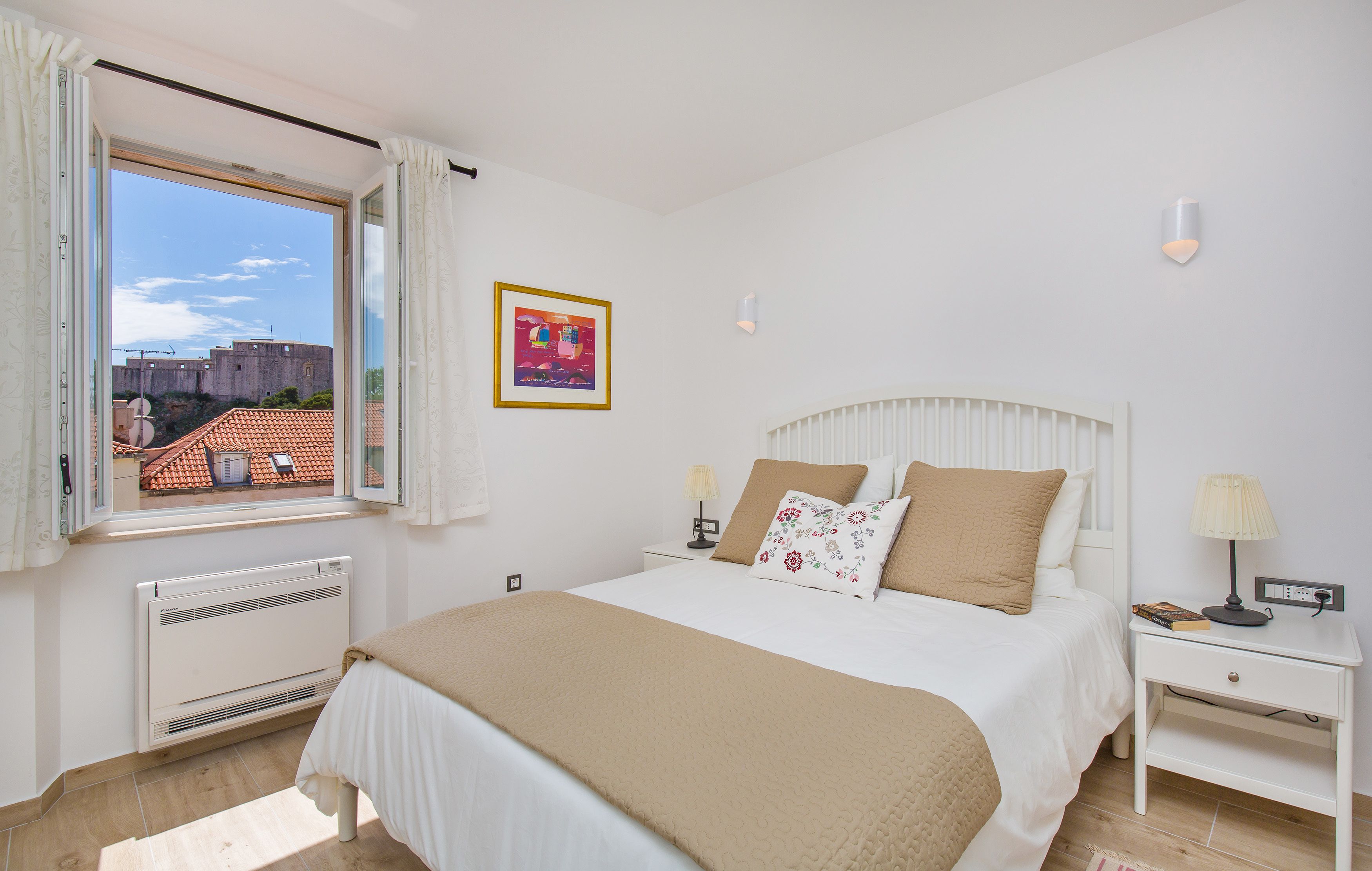 Ein Schlafzimmer mit Bett, Nachttischen, einem Fenster mit Vorhängen, einer Heizung und einem gerahmten Bild an der Wand.