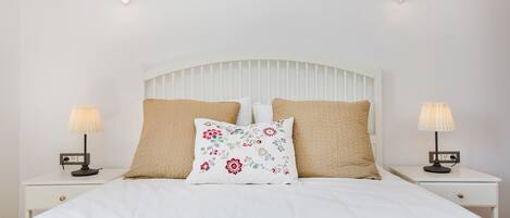Un lit bien fait, avec une tête de lit blanche, deux lampes de chevet et un coussin à motifs floraux.
