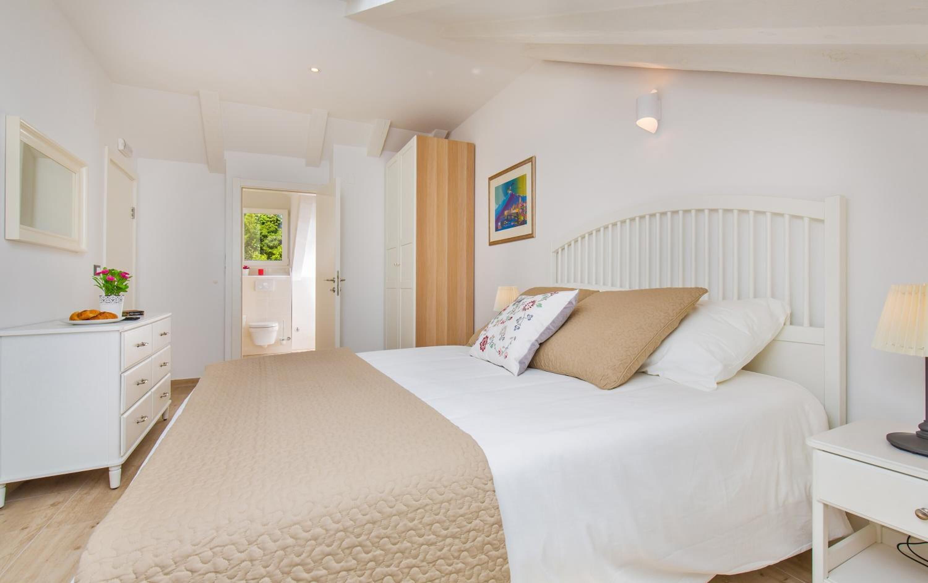 A bedroom with a bed, bedside table, and a dresser. There is a window with a view of greenery outside.