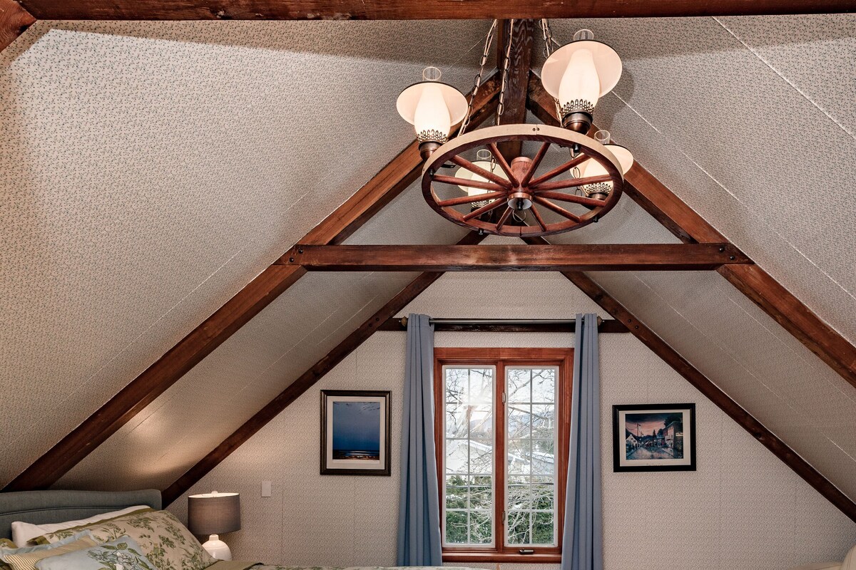 Ein Schlafzimmer mit schrägem Dach, Holzbalken, einem Bett mit floralem Bettzeug, einem Nachttisch, einer Kommode, einem Fenster mit Vorhängen und einer Deckenleuchte.