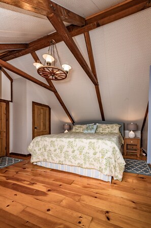 Ein Schlafzimmer mit Holzbalkendecke, einem Bett mit Blumenmuster-Bettwäsche, einem Nachttisch aus Holz und einer Pendelleuchte.