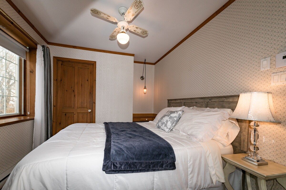 Ein Schlafzimmer mit Holzakustikdeckenventilator, einem Bett mit weißer Bettwäsche, einem Nachttisch mit Lampe und einem Fenster mit Vorhängen.