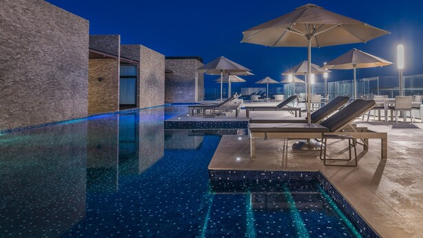 A nighttime swimming pool area with lounge chairs and umbrellas, surrounded by a modern building with a brick facade. - New and modern 2BR downtown apartment by Happy Address (Playa del Carmen)