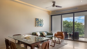 Un comedor moderno con una mesa de vidrio, sillas de madera, un sofá verde y un amplio ventanal con vistas a la vegetación.