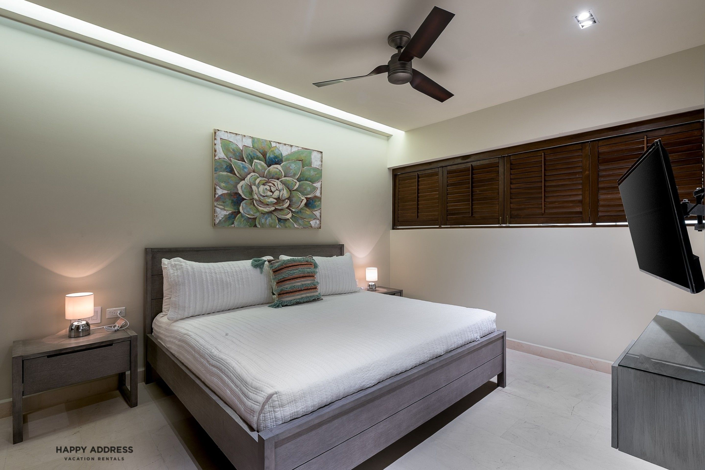 A bedroom with a large bed, a ceiling fan, a wall-mounted TV, and a window with wooden shutters.
