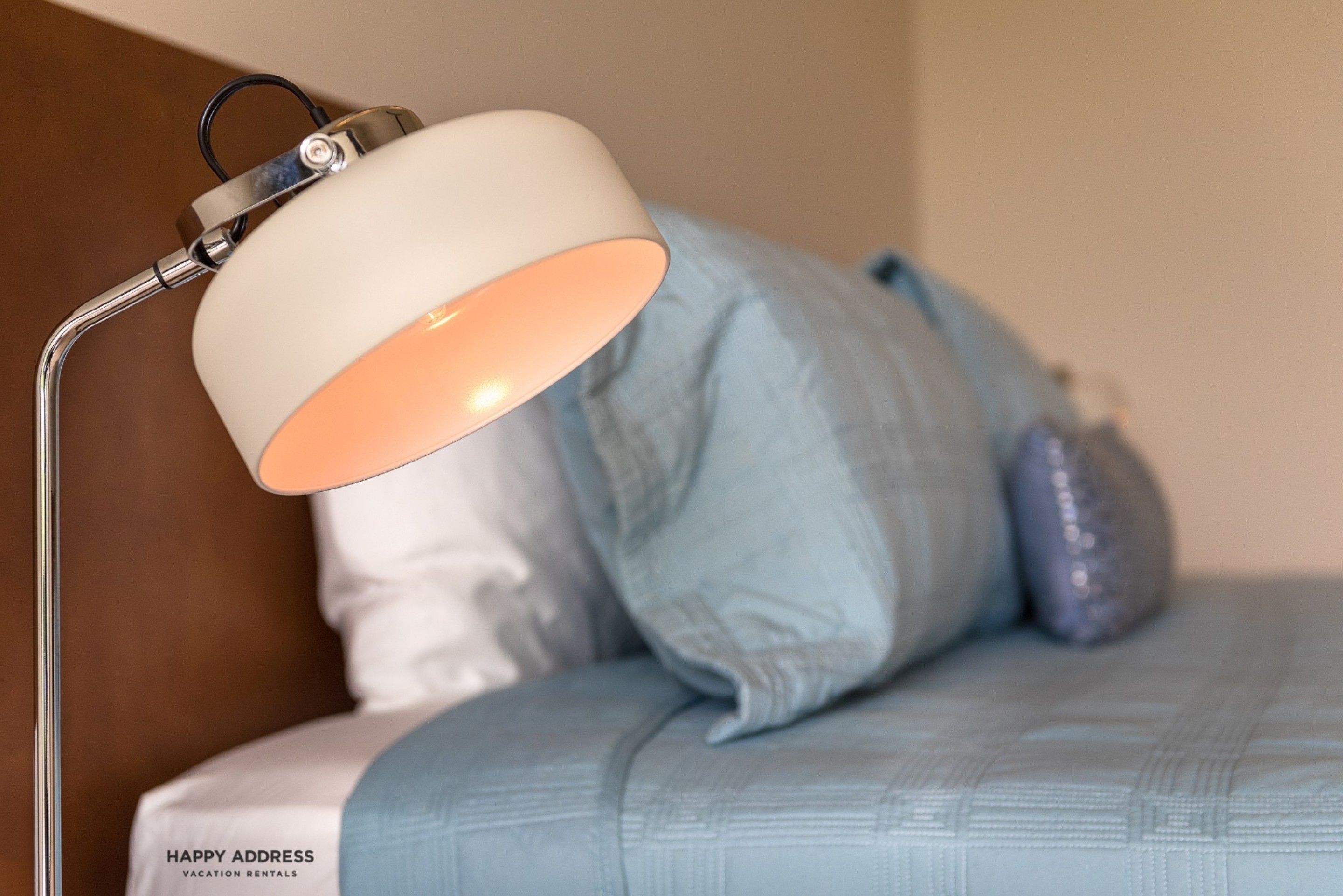 Ein Bett mit blauer Bettdecke und Kissen, ein weißes Kissen mit Text sowie eine Lampe mit weißem Lampenschirm.