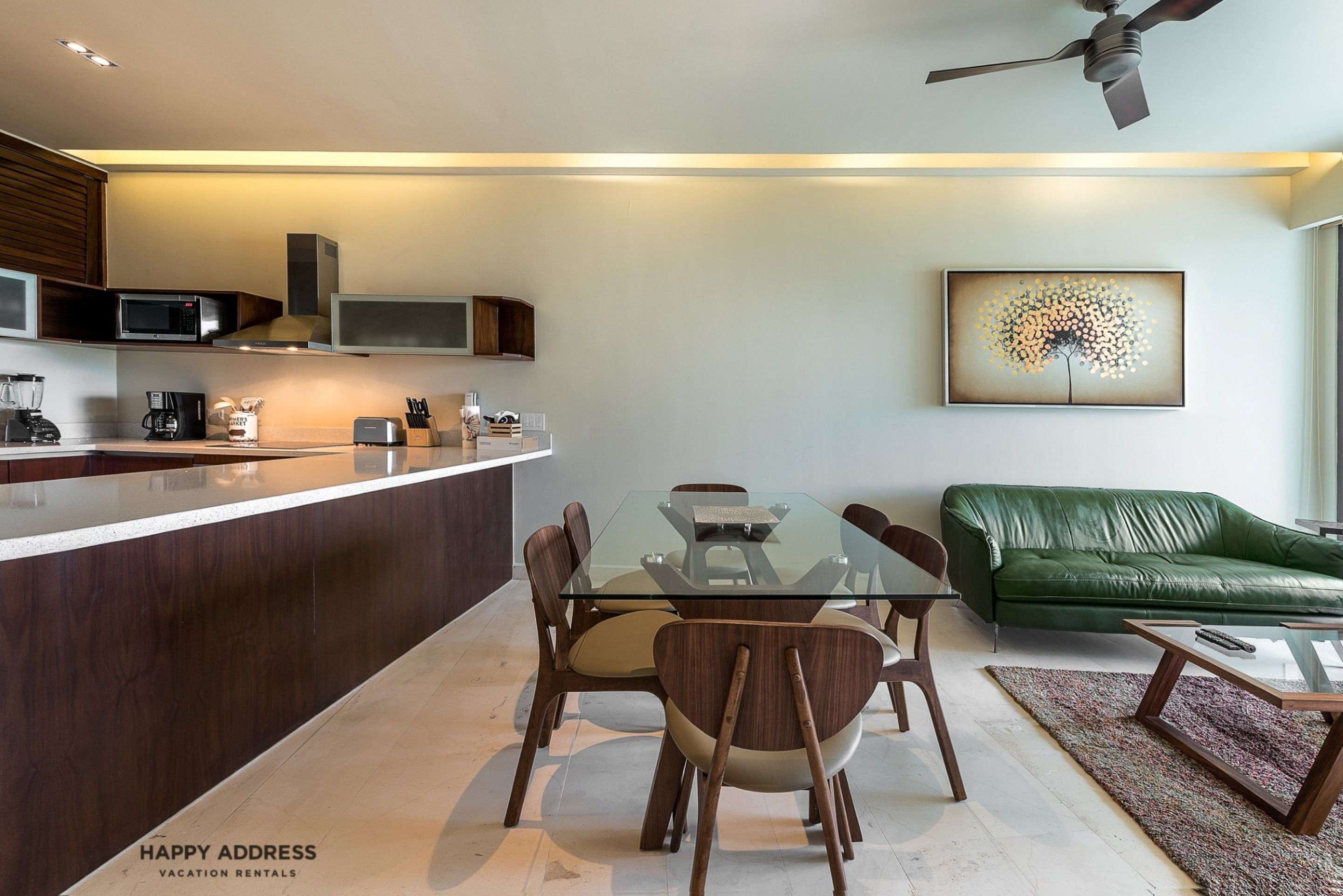 A modern kitchen with a glass dining table, wooden chairs, a green leather sofa, and a ceiling fan.