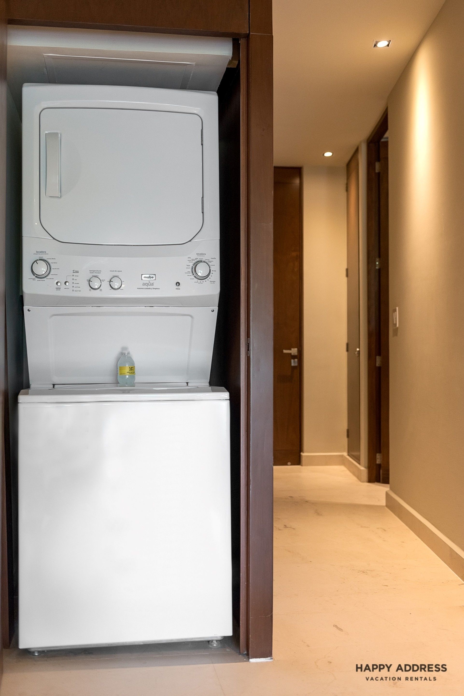 A compact laundry room with a front-loading washer and dryer, a bottle of hand soap, and a hallway leading to a door.