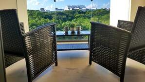 A balcony with wicker chairs and a glass table, overlooking a lush green landscape and city buildings. - Pleasant 1BR a few step away from the beach by Happy Address (Playa del Carmen)