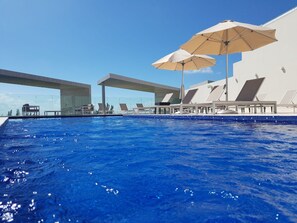 A swimming pool with lounge chairs and umbrellas, surrounded by a modern building with a glass facade. - Brand New 1 BR Condo with jungle view by Happy Address (Playa del Carmen)