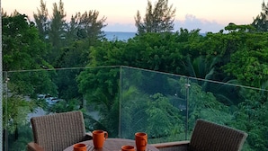 Un balcon agrémenté de meubles en osier et de tasses orange, offrant une vue sur un paysage verdoyant et luxuriant ainsi que sur une étendue d’eau.