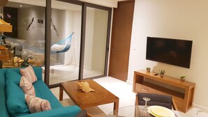 A modern living room with a teal sofa, a wooden coffee table, a glass dining table set for two, and a large flat-screen TV mounted on the wall. - Caribbean Lifestyle 2BR steps from the beach by Happy Address (Playa del Carmen)