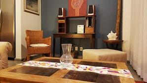 Un soggiorno con un tavolo in legno, una poltrona in vimini, un tavolino laterale con un oggetto decorativo e una libreria con diffusori.