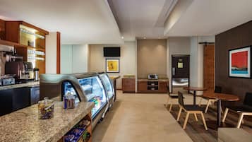 A hotel lobby with a self-service coffee station, a refrigerated display case, seating area, and a television mounted on the wall.