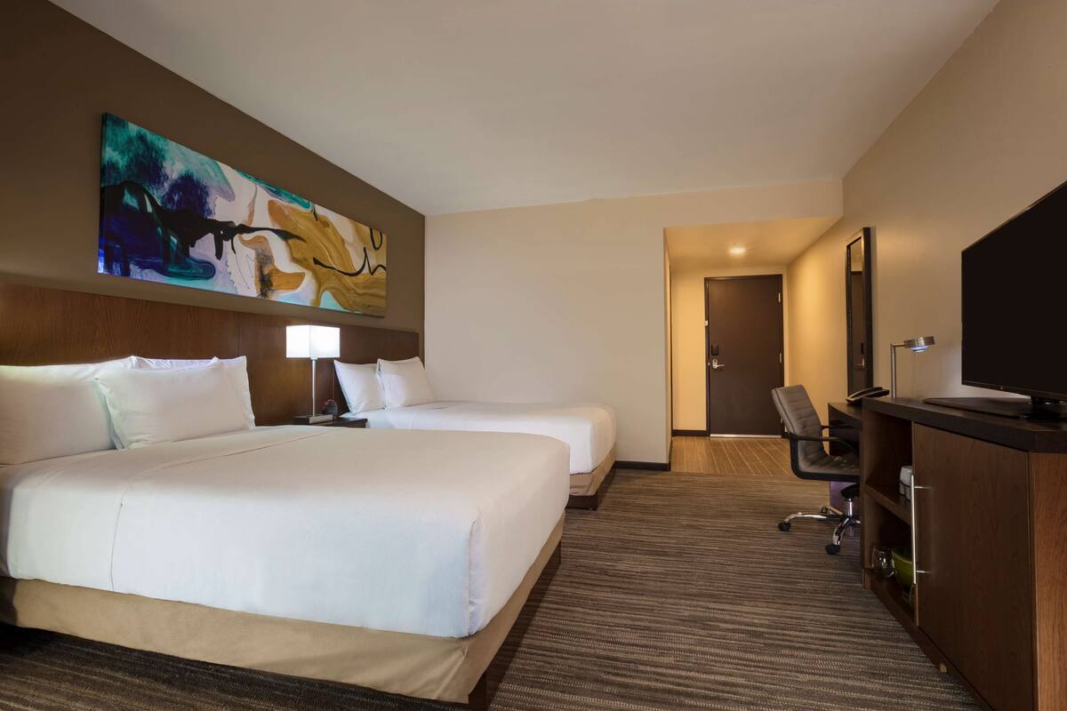 A hotel room with two beds, a desk, a chair, a television, and a painting on the wall.